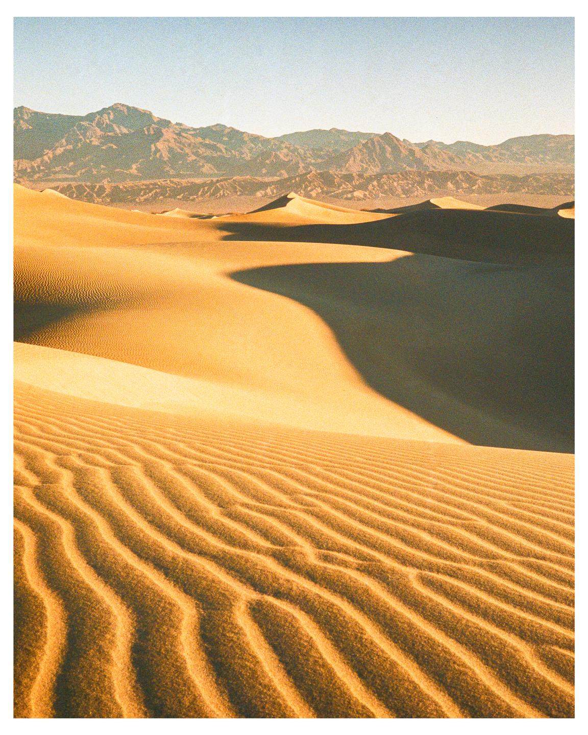 A desert landscape shows sand dunes with ripples and shadows, and mountains in the distance under a clear sky.