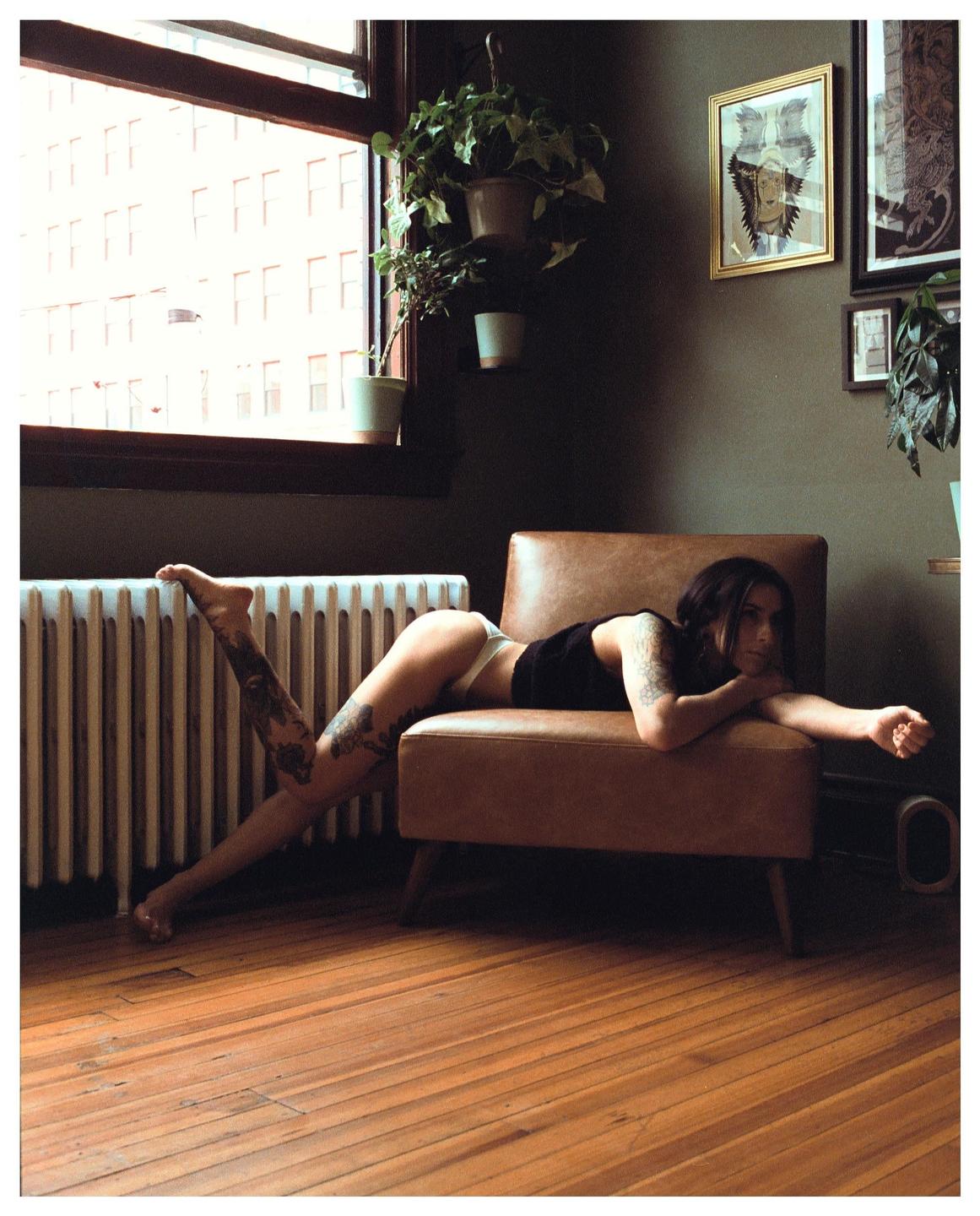 A woman with tattoos is lying on a brown armchair next to a window with potted plants.