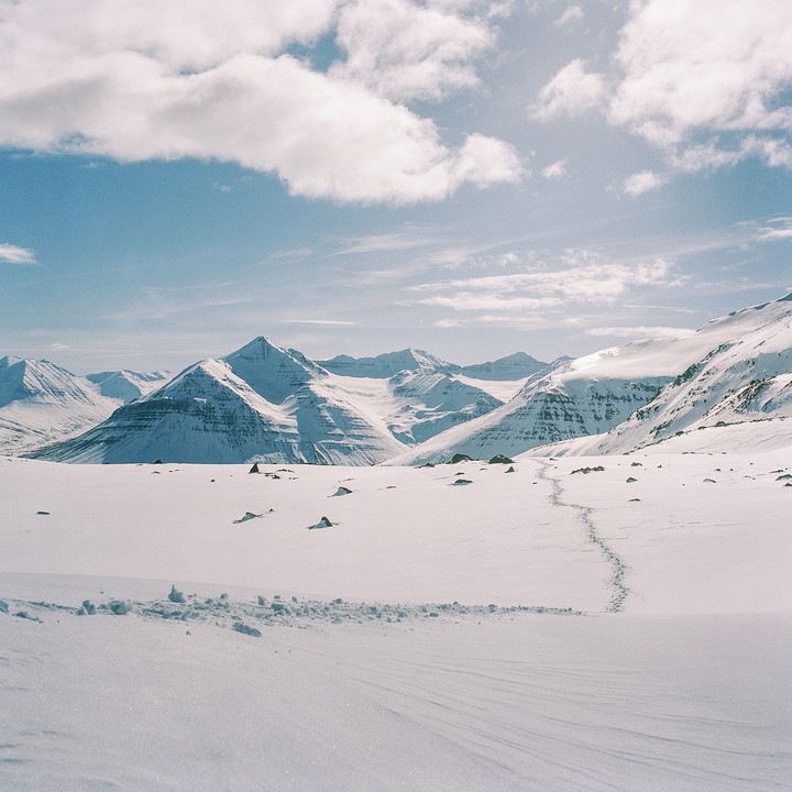 A vast, snow-covered landscape stretches towards a range of mountains under a bright sky with clouds.