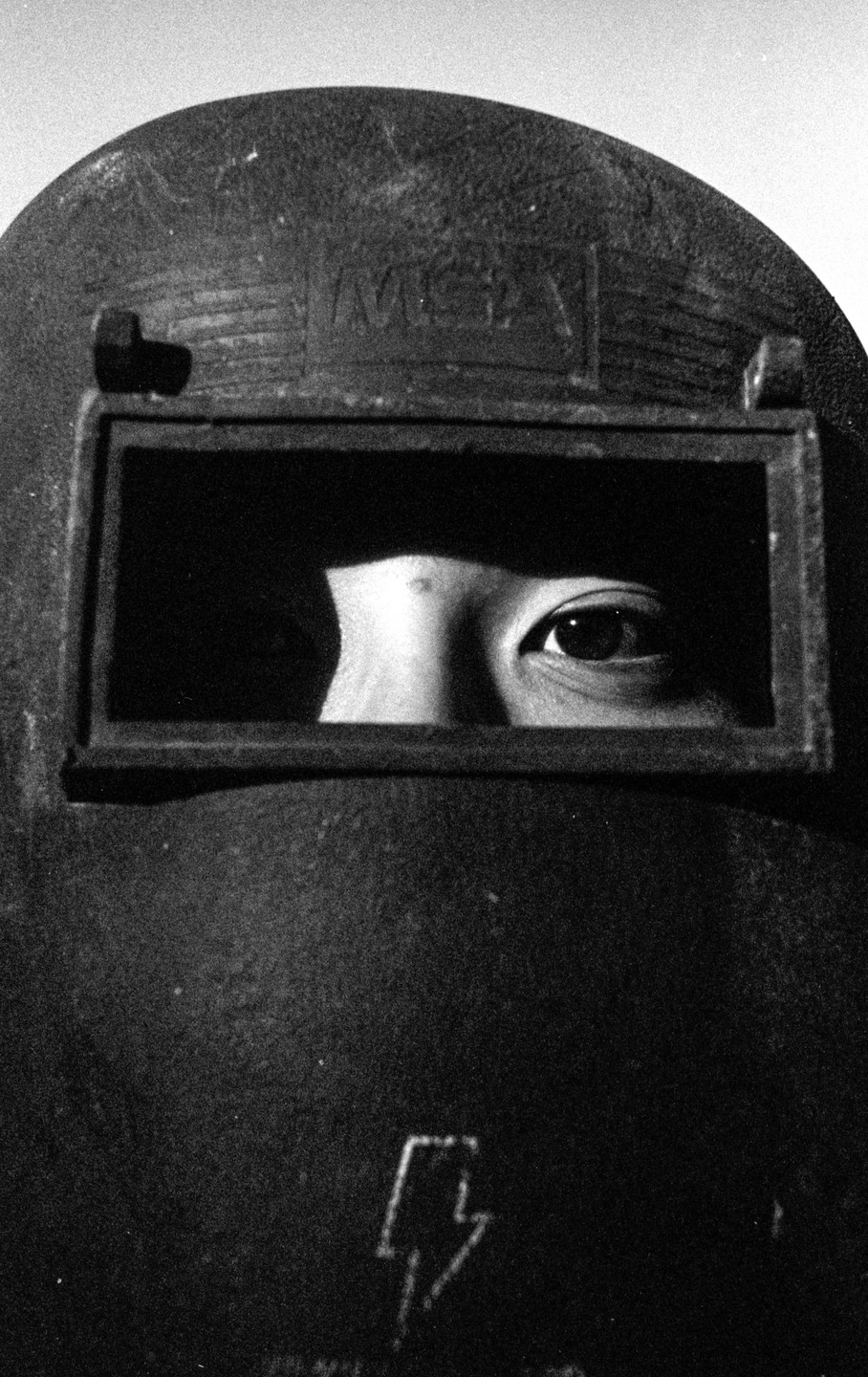A person's eye is visible through the rectangular opening of a dark welding helmet.