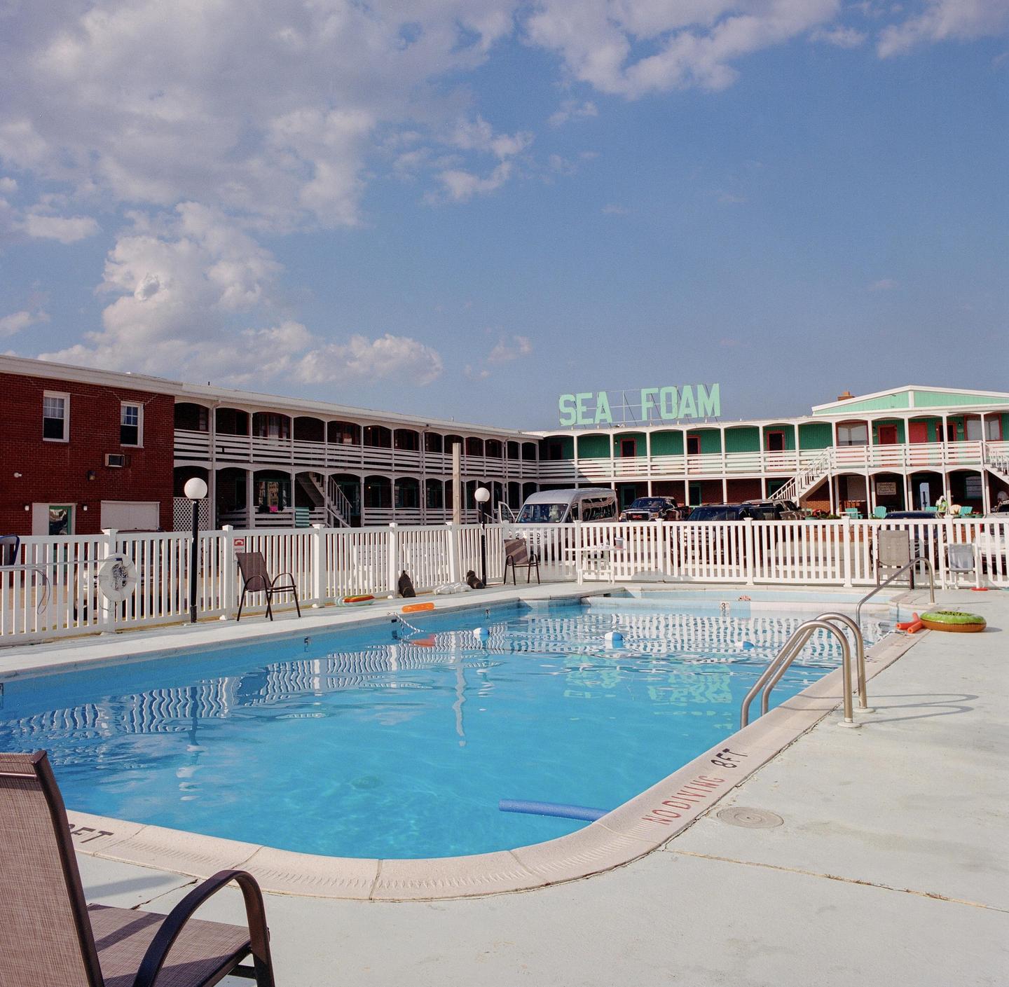 A swimming pool with a white fence is in front of a motel with a "SEA FOAM" sign on the roof.