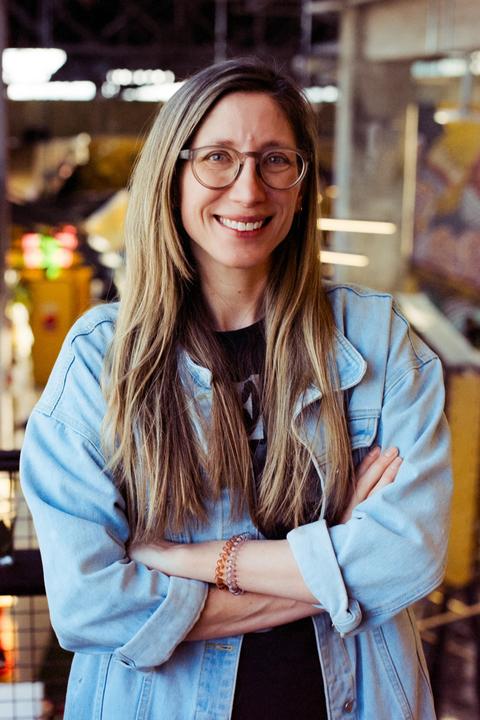 A woman with long hair and glasses smiles at the camera, wearing a denim jacket with her arms crossed.