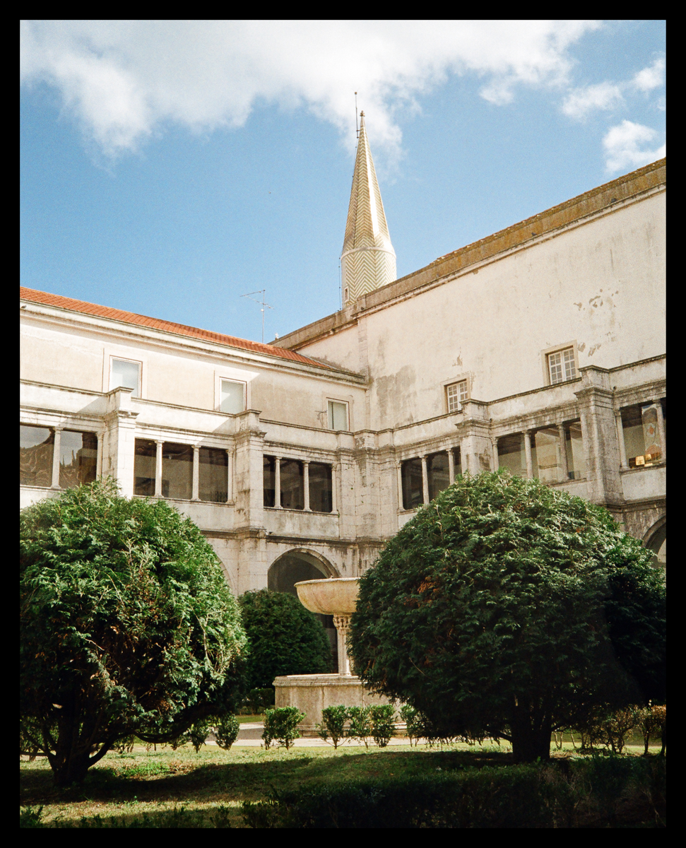 A courtyard with a fountain and manicured bushes is surrounded by a building with a tall, pointed spire.