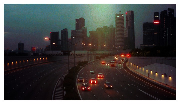 A highway curves through a city with tall buildings and car lights visible at dusk.