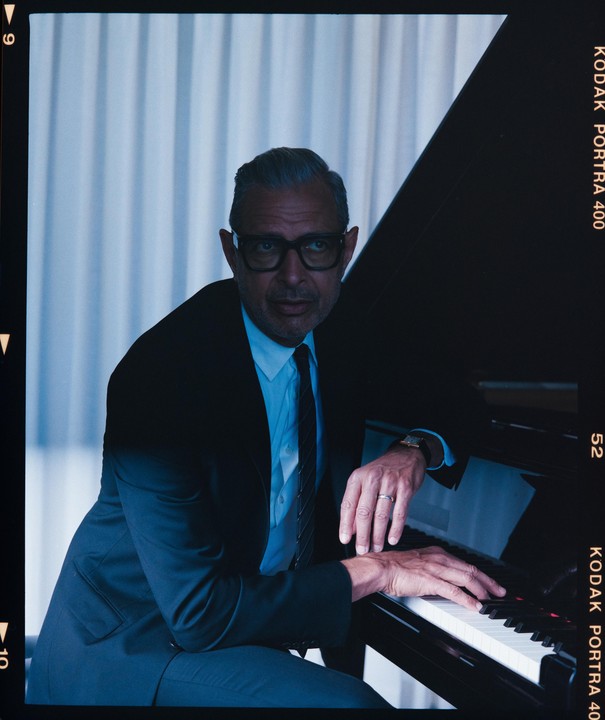 A man in a suit and glasses sits at a piano, looking towards the viewer.