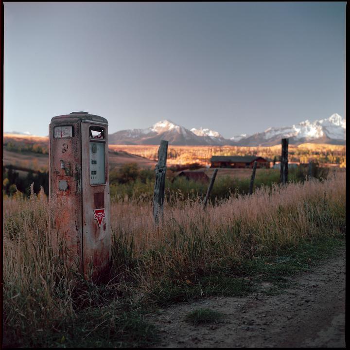 An old gas pump stands in a field of tall grass with mountains in the background.