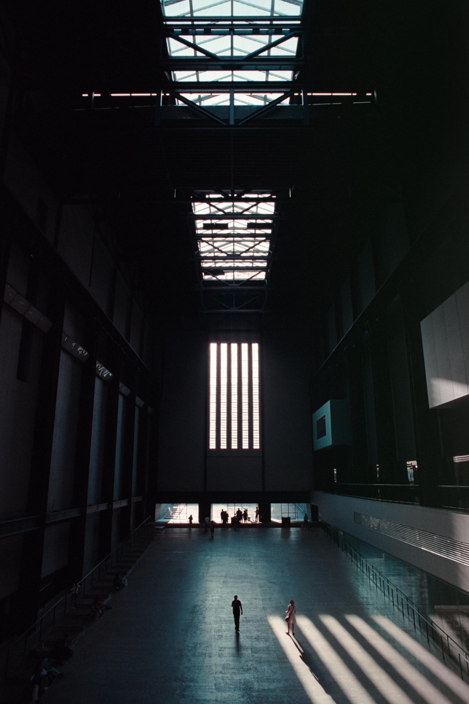 Two people walk across a large, dark hall with light streaming in from high windows and skylights.