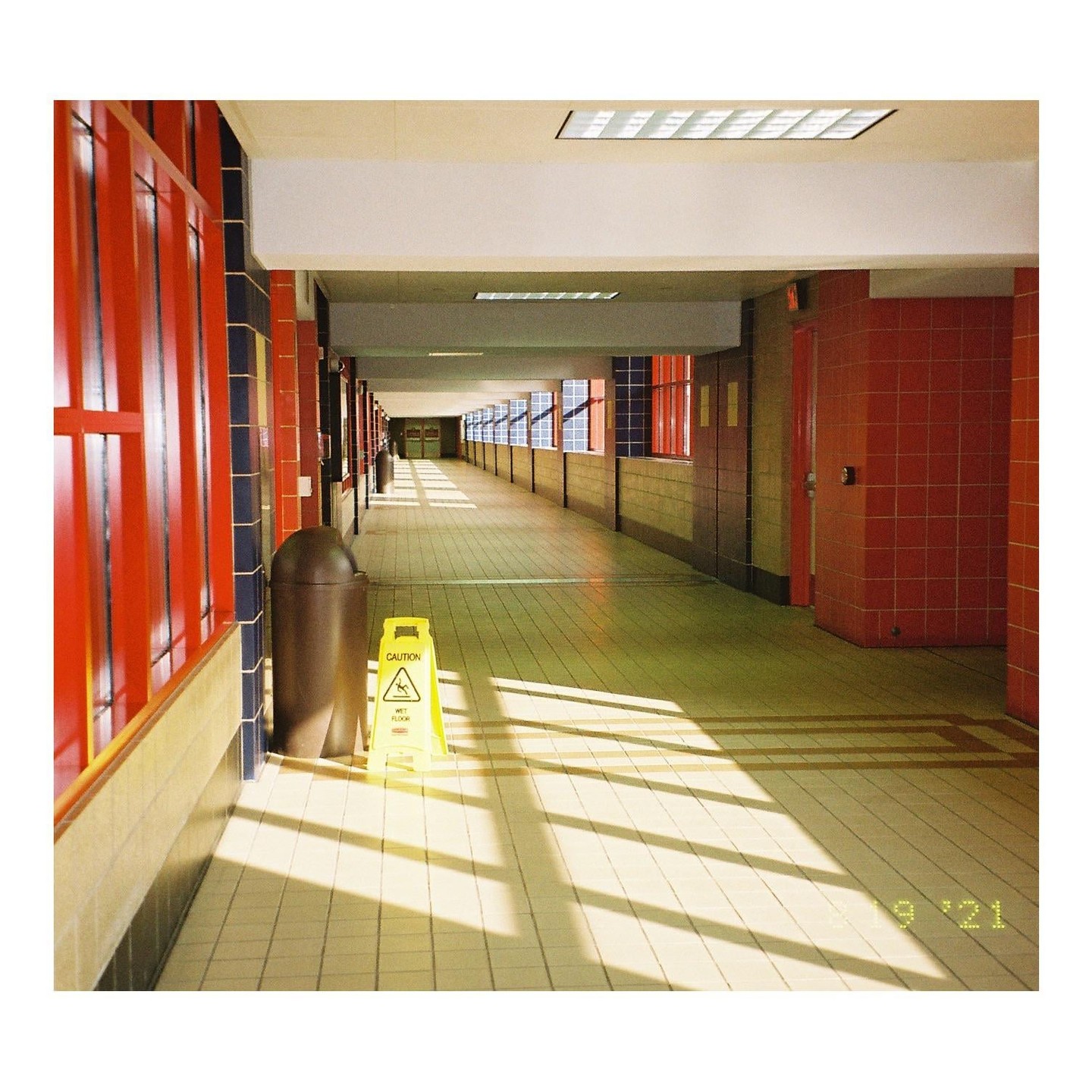 A long, empty hallway with red and blue walls is illuminated by sunlight streaming through windows, casting shadows on the tiled floor where a wet floor sign stands next to a trash can.