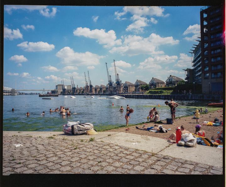 People swim and relax on the shore of a river with cranes and buildings in the background.