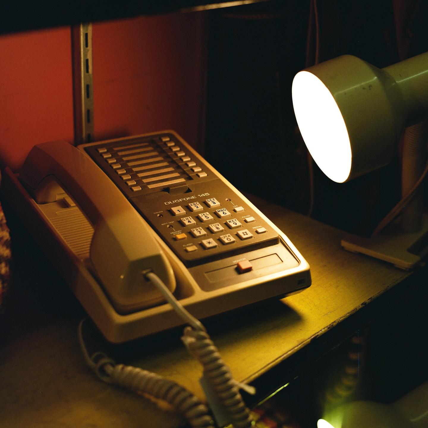 A landline telephone and a desk lamp are on a shelf in a dimly lit room.
