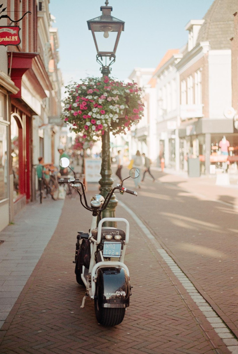 A white scooter is parked on a brick sidewalk next to a lamppost adorned with a hanging basket of pink and white flowers, with a street and buildings in the background.