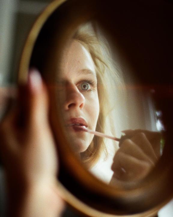 A woman applies lip gloss while looking into a mirror.