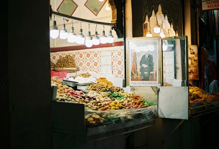 A brightly lit stall in a market displays a variety of colorful pastries and a framed portrait of a man.