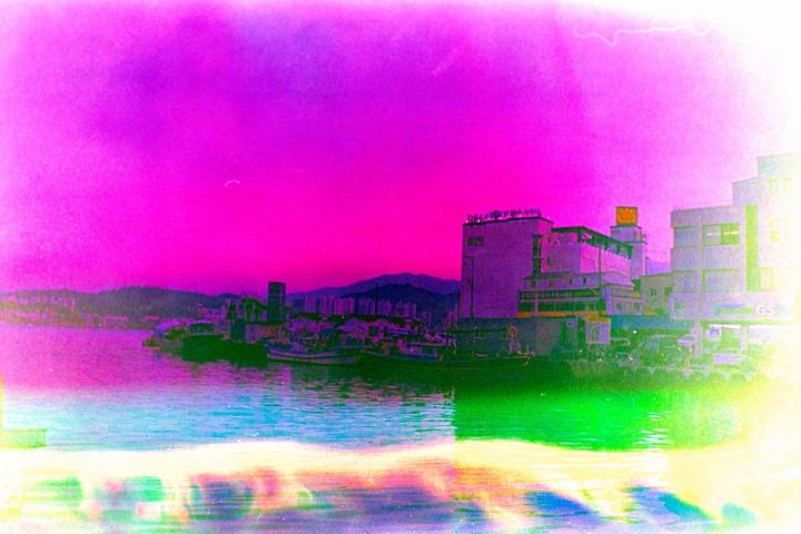A vibrant, color-shifted image shows a body of water with boats and buildings along the shore under a bright pink sky.