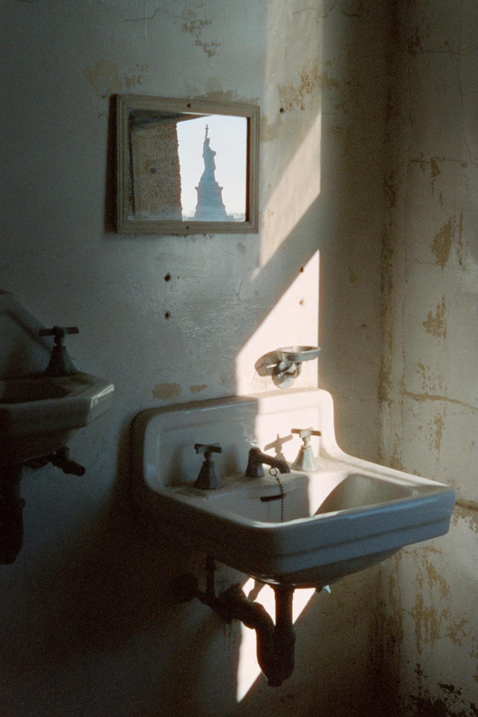 Sunlight illuminates two sinks in a dilapidated room, with the Statue of Liberty visible in a small mirror on the wall.