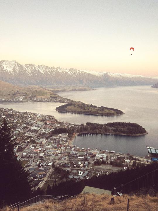 A paraglider flies over a town next to a lake with snow-capped mountains in the background.