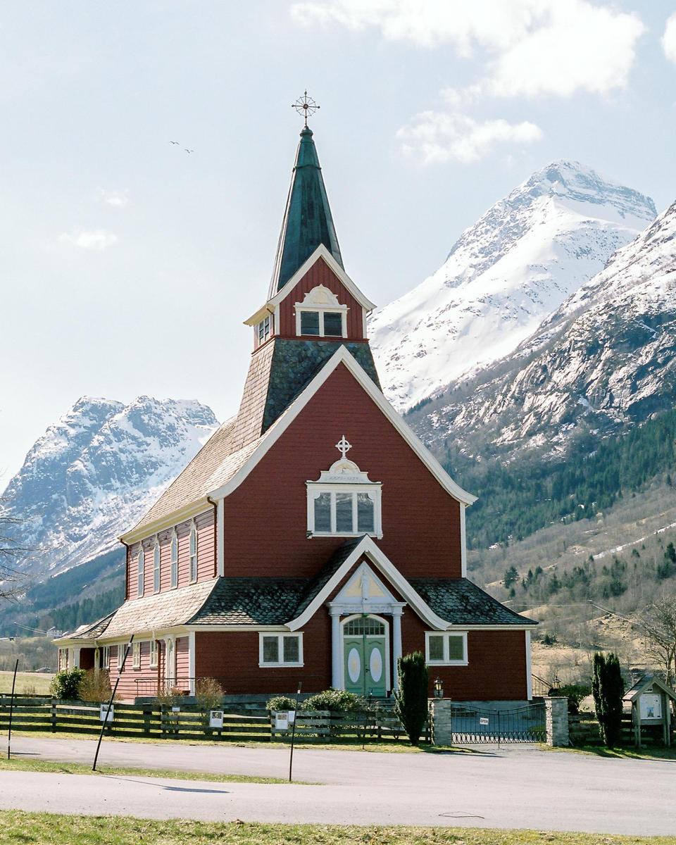 A red church with a tall steeple stands in front of snow-capped mountains.