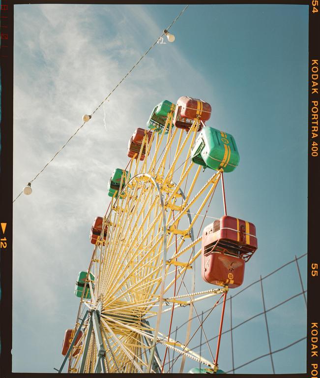 A low-angle view shows a colorful ferris wheel against a blue sky with string lights hanging in the foreground.