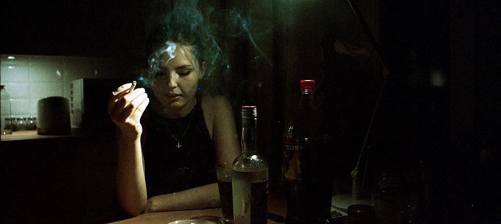 A woman sits at a table in a dark room, holding a lit cigarette with smoke rising around her head.