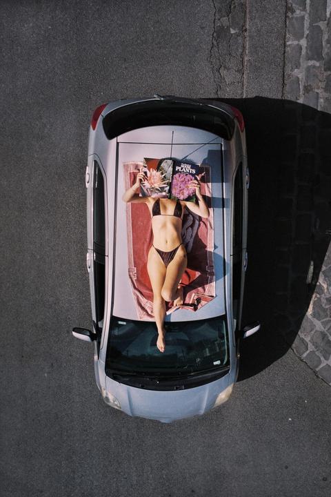 A woman in a bikini lies on a towel on the roof of a car, holding a book with flowers on the cover over her face.