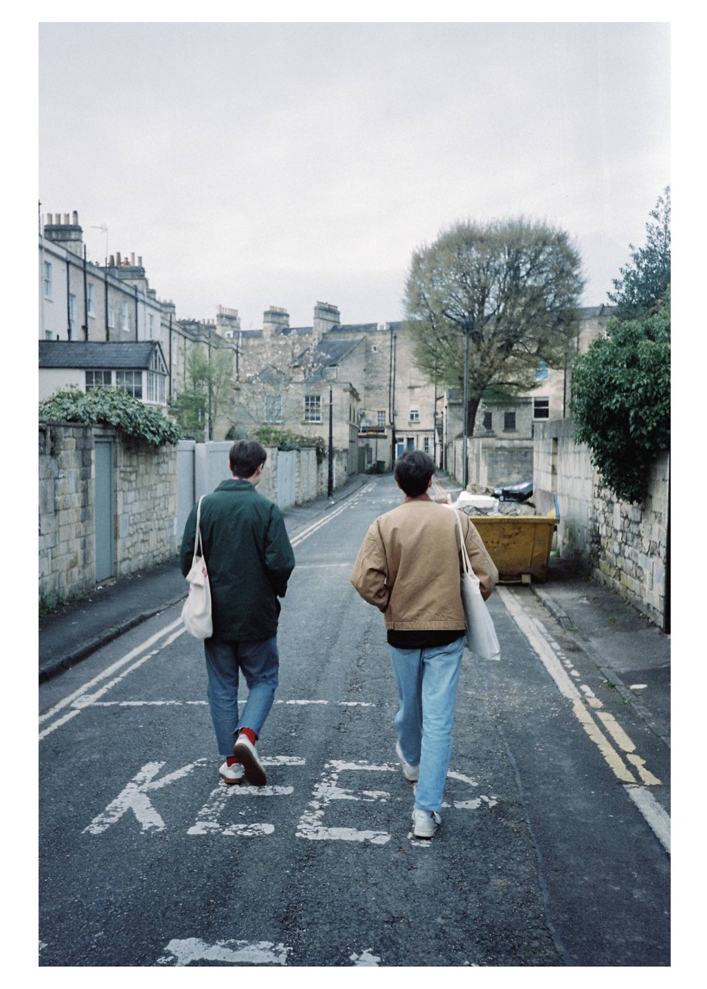 Two men walk away from the viewer down a street with stone buildings and a large tree.