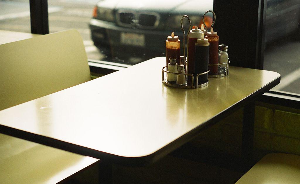 A diner booth table with a condiment caddy sits next to a window looking out onto a street with a car.