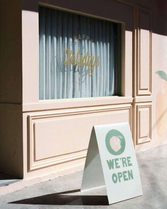A white A-frame sign with green text that reads "WE'RE OPEN" stands in front of a peach-colored building with a window displaying "Happy Holidays" in gold lettering.