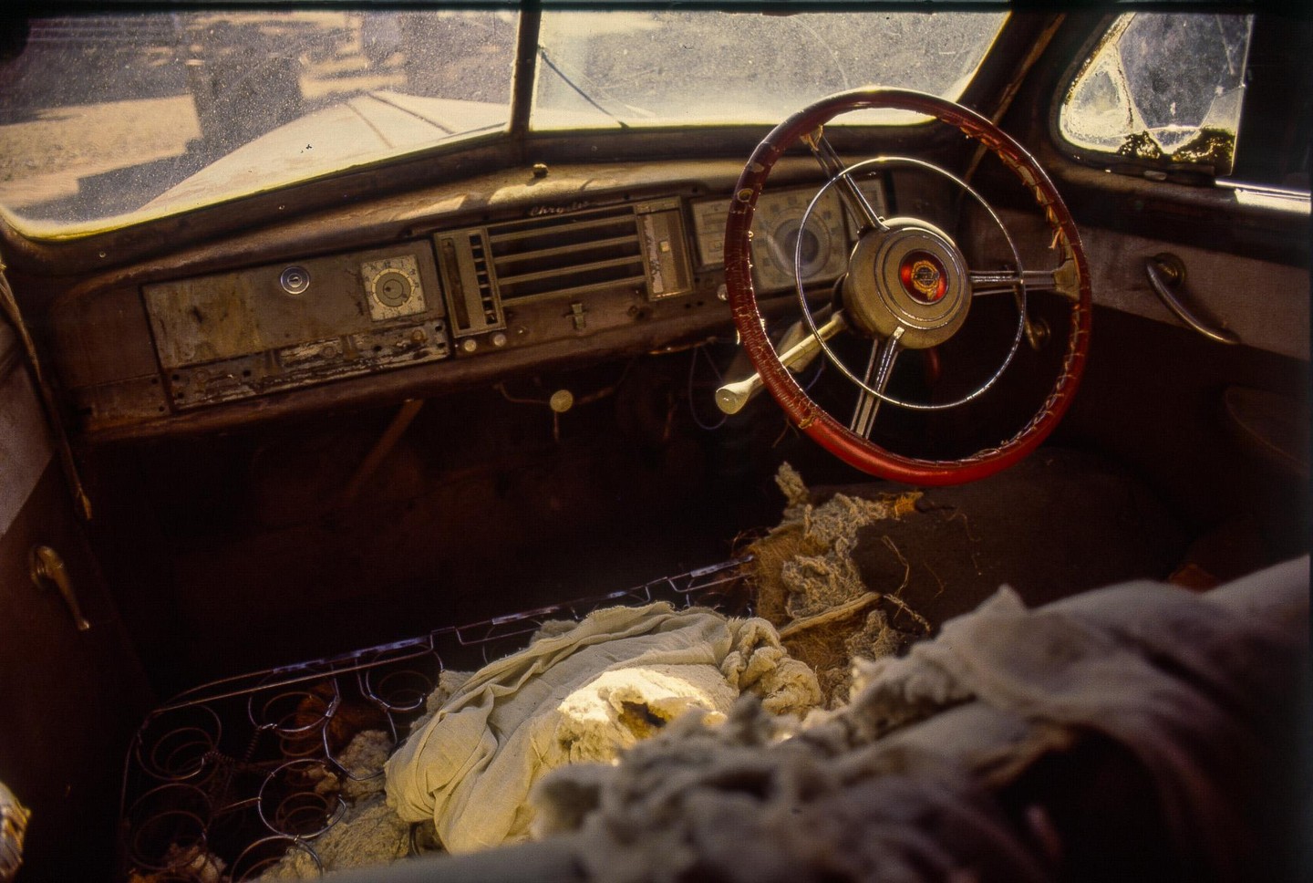 The interior of an old car shows a red steering wheel, dashboard, and torn upholstery.
