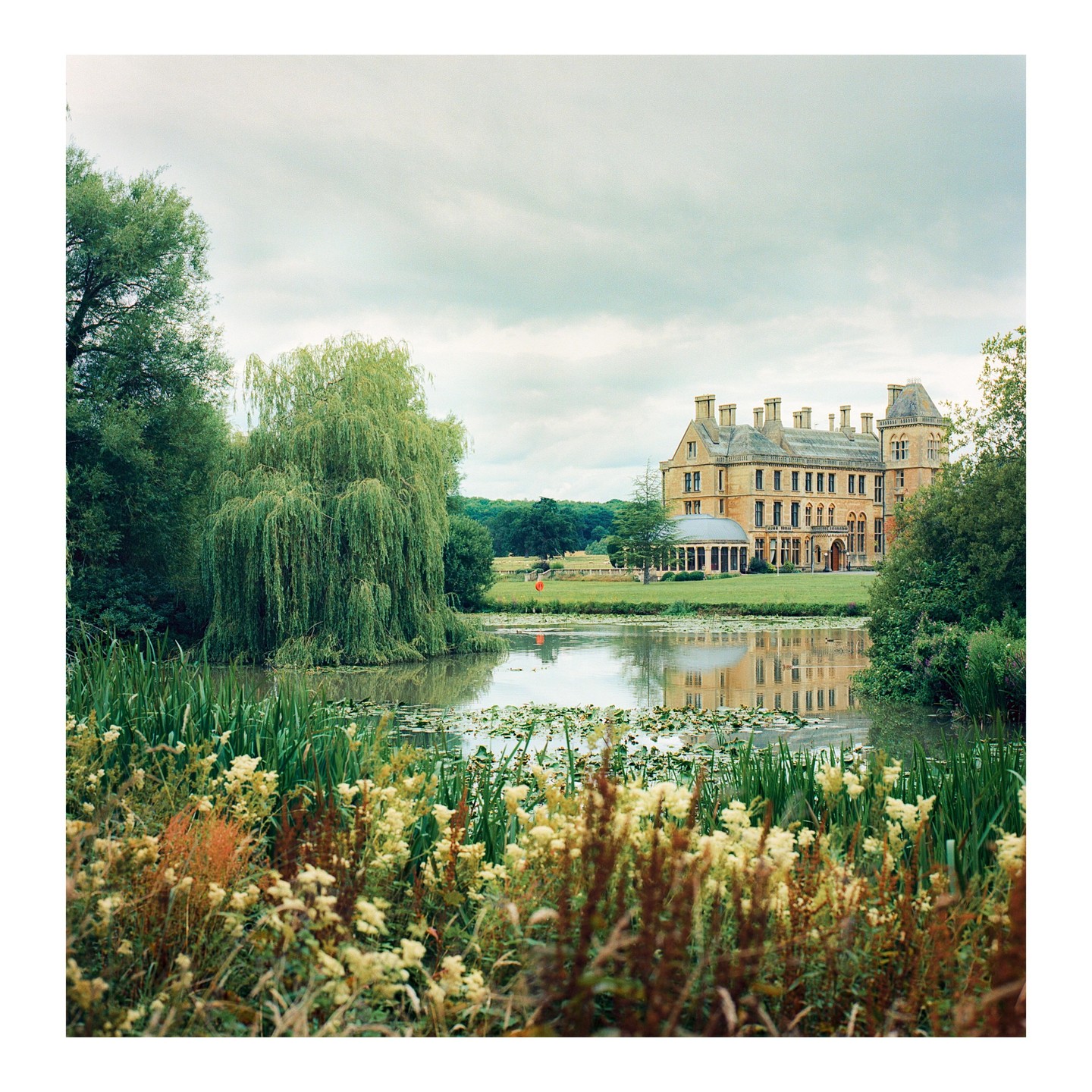 A large manor house is reflected in a pond surrounded by lush vegetation and a weeping willow tree.
