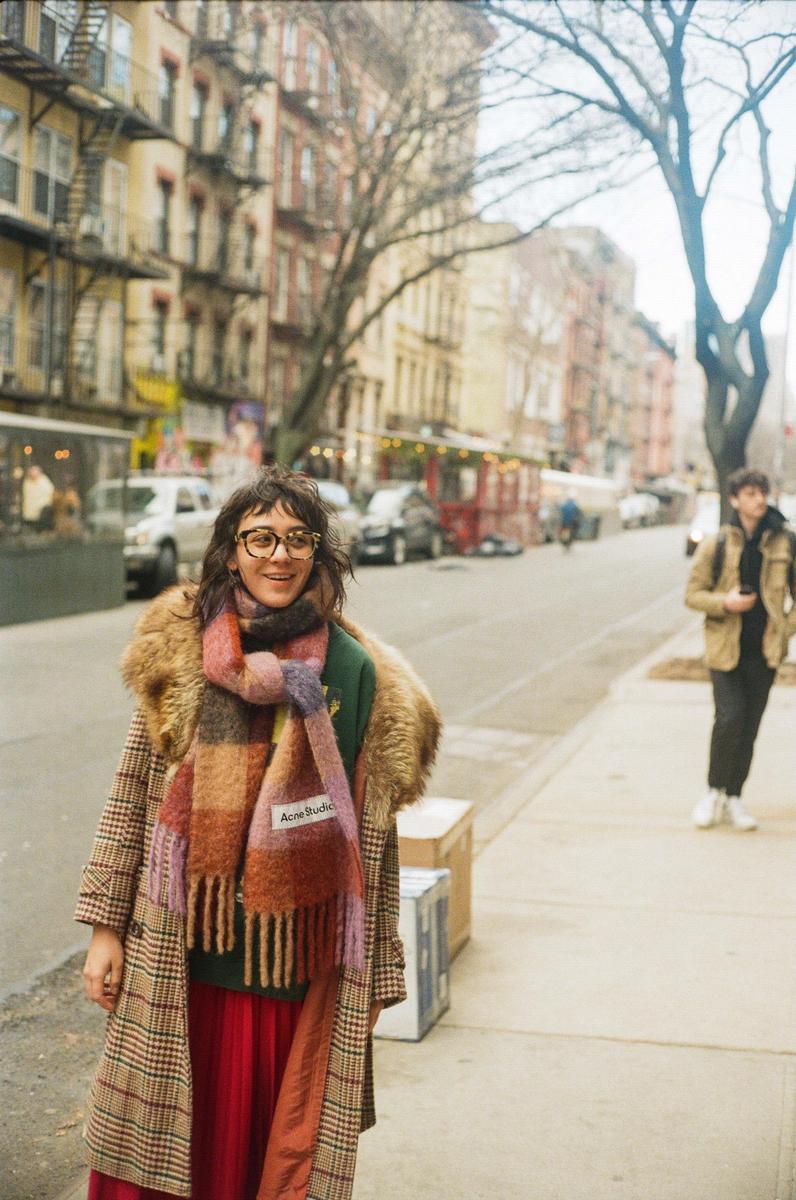 A woman wearing a plaid coat and a colorful scarf smiles at the camera on a city street.