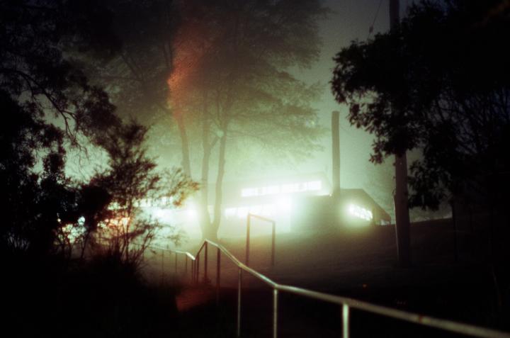 A foggy night scene shows a building with illuminated windows and a handrail leading down a path, surrounded by trees.