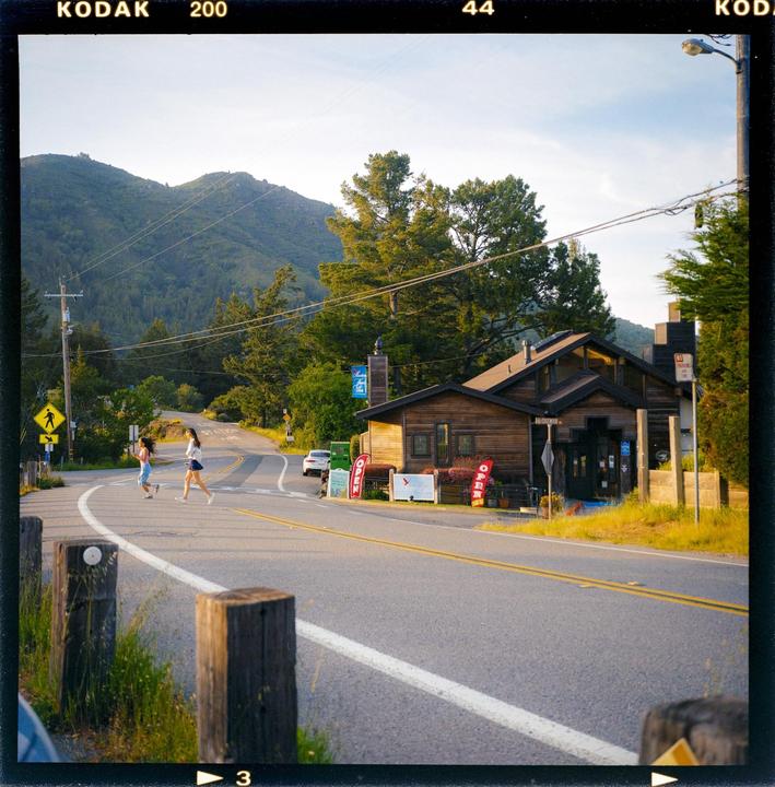 Two women cross a winding road in front of a wooden building with mountains and trees in the background.
