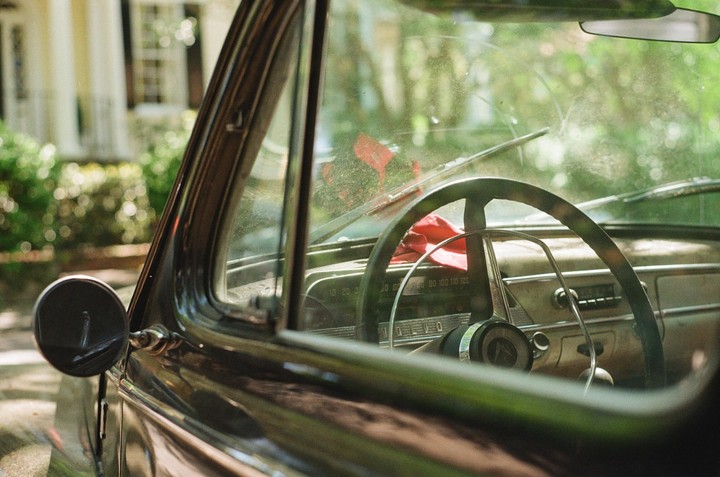 The interior of a vintage car is visible through the dusty windshield, with a red cloth draped over the steering wheel.