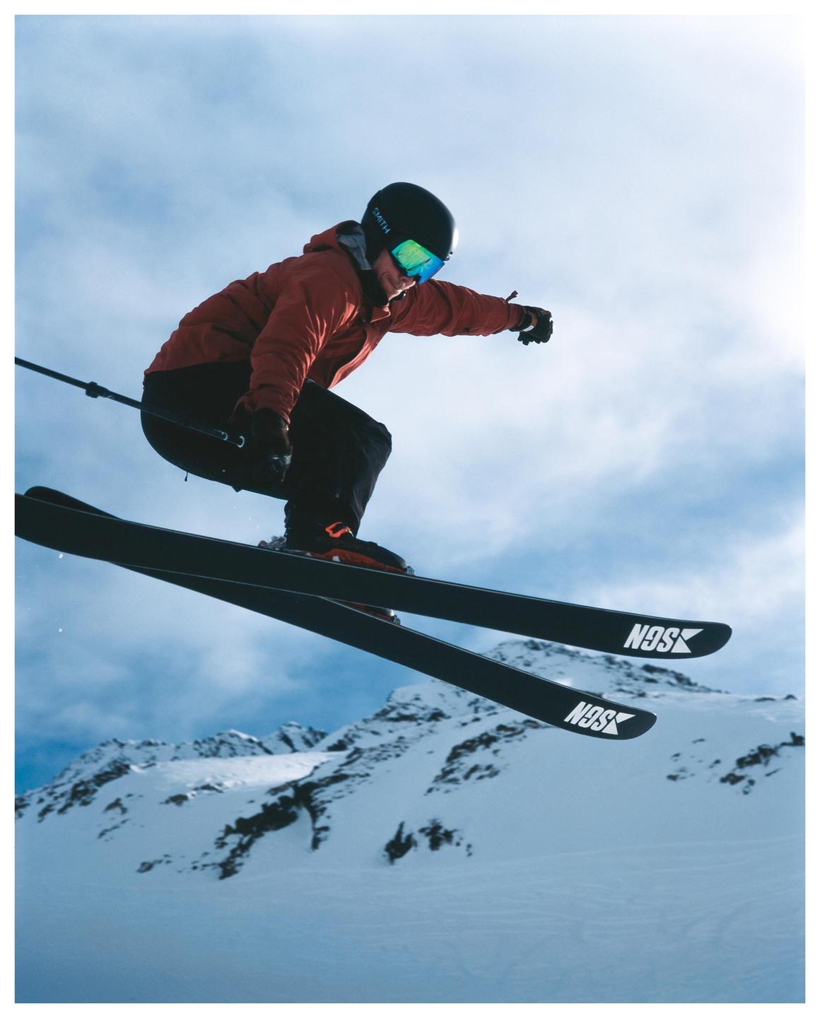 A skier in a red jacket and black helmet is captured mid-air against a backdrop of snow-covered mountains and a cloudy sky.
