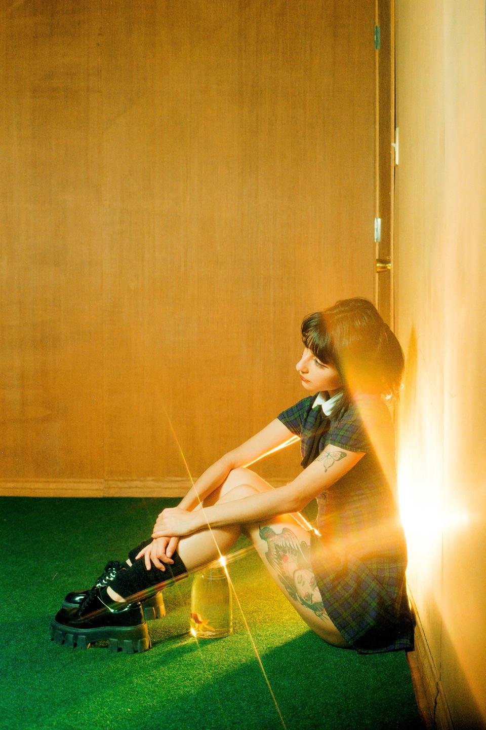 A woman sits on a green carpet next to a wall with a fish in a jar beside her.