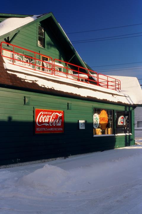 A green building with a red Coca-Cola sign and a Shell sign in the window is covered in snow under a clear blue sky.