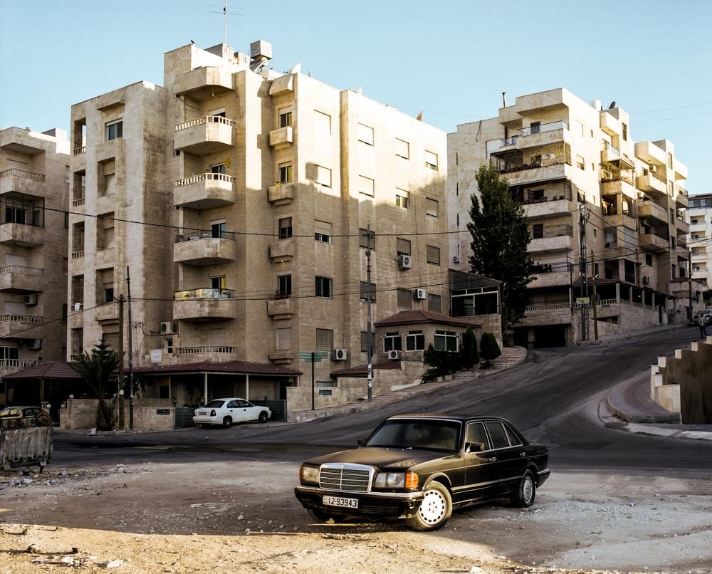 A black car is parked on a dirt patch next to a street with large buildings in the background.