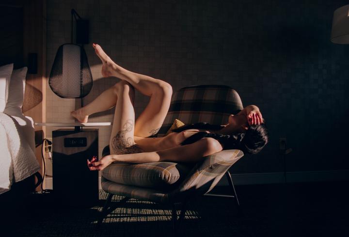 A woman reclines on a plaid armchair with her legs up and her arm over her face, illuminated by a shaft of light.