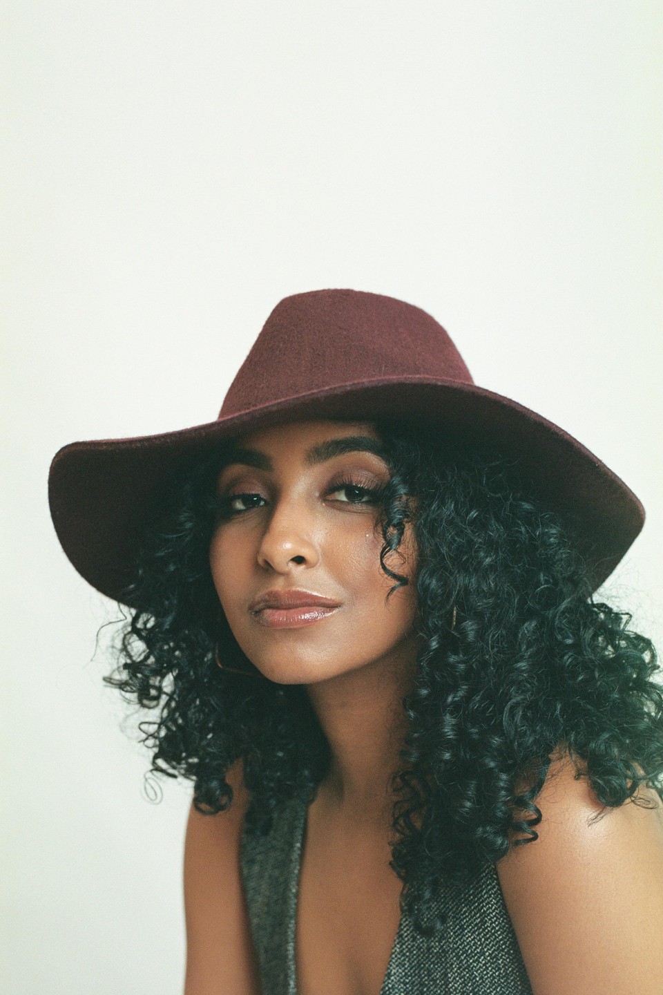 A woman with curly hair and a wide-brimmed hat looks directly at the viewer.