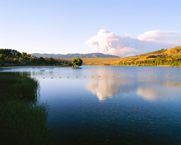 A lake with a line of buoys is surrounded by hills and trees under a sky with a large cloud.