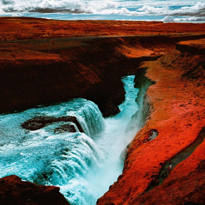 A waterfall cascades into a canyon with vibrant orange foliage on the surrounding cliffs under a cloudy sky.
