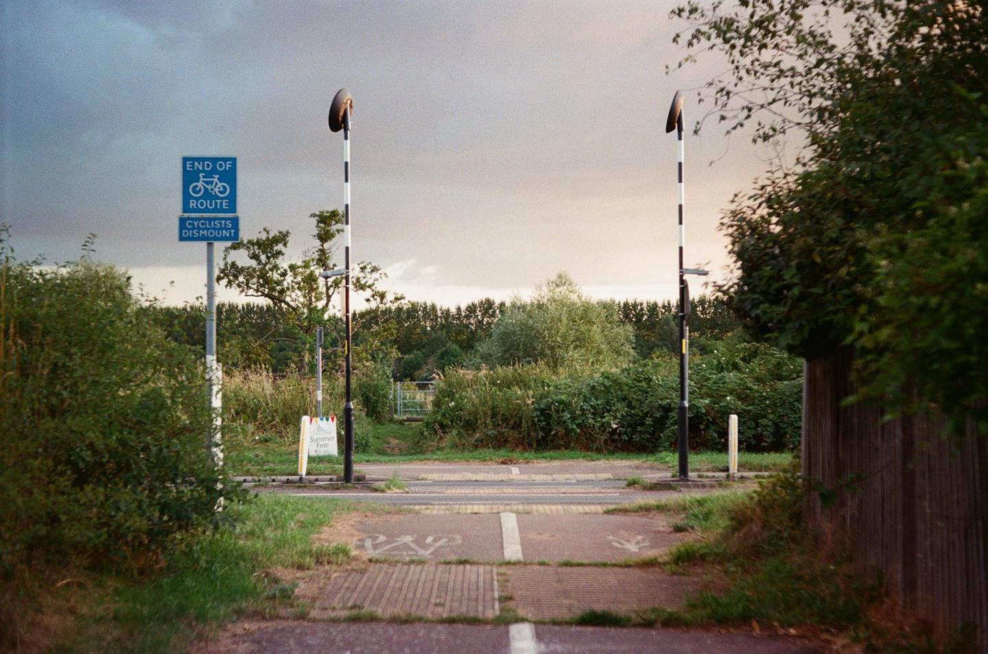 A bicycle route sign stands next to a path with two striped poles and a wooden fence.