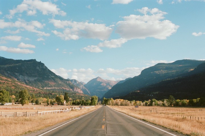 A long road stretches through a valley with mountains on either side under a blue sky with clouds.
