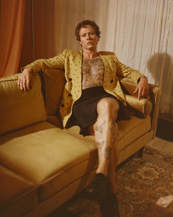 A man with a tattooed chest and legs sits on a yellow sofa, wearing a yellow jacket and a black skirt.