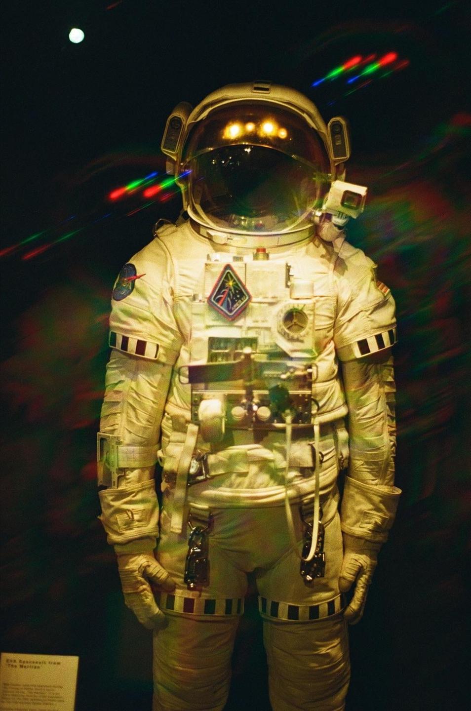 An astronaut suit stands in a dark room with colorful light streaks and a bright orb in the background.
