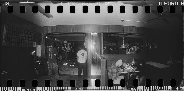 A band performs on a stage in a dark bar while people watch and sit at tables.