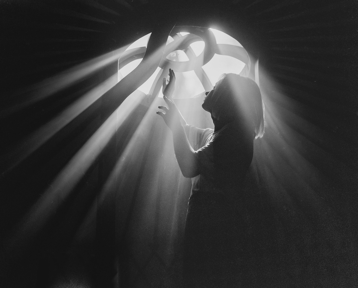 A woman stands in a dark room with her hands raised, illuminated by beams of light streaming through a round window.