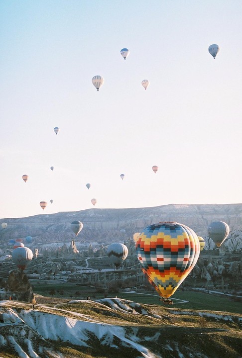 Numerous hot air balloons float above a valley with unique rock formations and green fields under a clear sky.