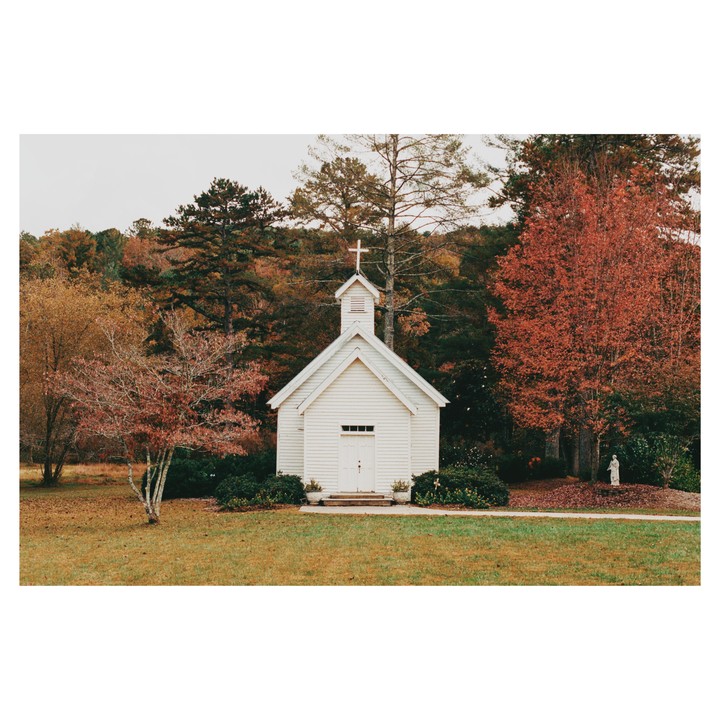 A small white church with a cross on its steeple stands in a grassy field surrounded by trees with autumn foliage.