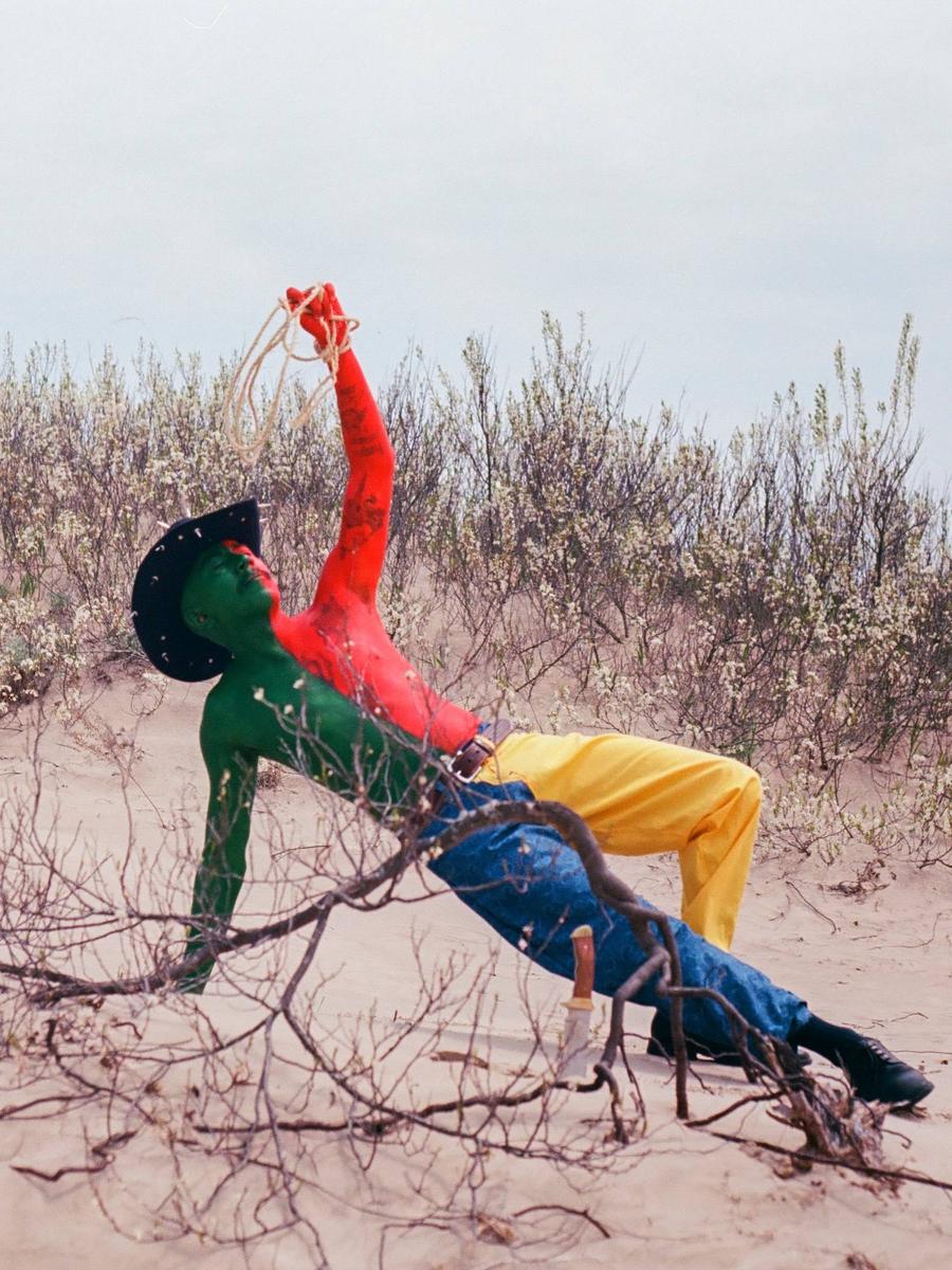 A person with red and green body paint and a cowboy hat holds a rope while leaning back in a desert landscape.